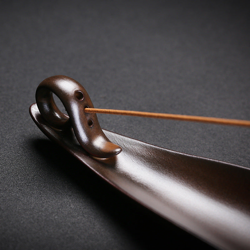 The Drift incense burner — leaf-shaped dark wood single-stick holder with incense burning gently against a dark background.