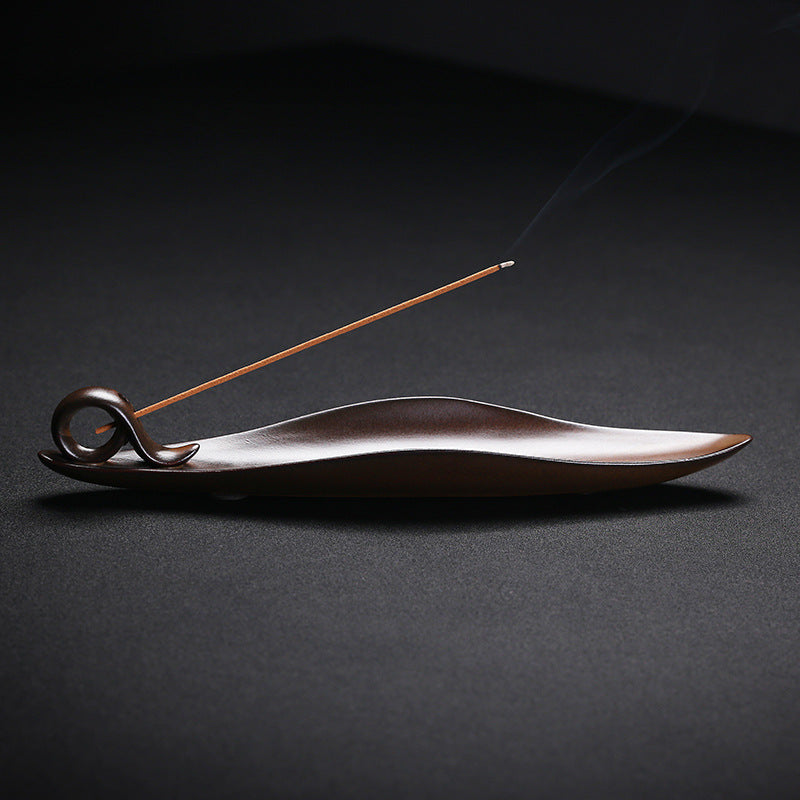 The Drift incense burner — leaf-shaped dark wood single-stick holder with incense burning gently against a dark background.