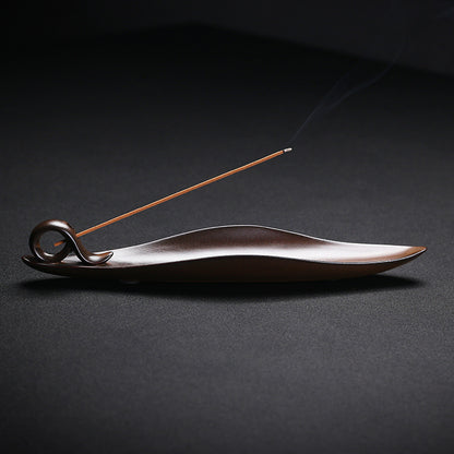 The Drift incense burner — leaf-shaped dark wood single-stick holder with incense burning gently against a dark background.