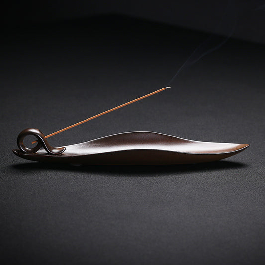 The Drift incense burner — leaf-shaped dark wood single-stick holder with incense burning gently against a dark background.